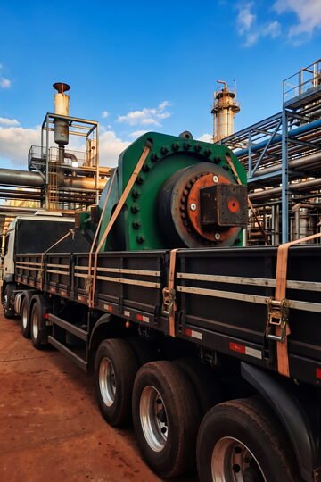 Equipamento industrial de grande porte amarrado sobre a carroceria, dentro de uma planta industrial.